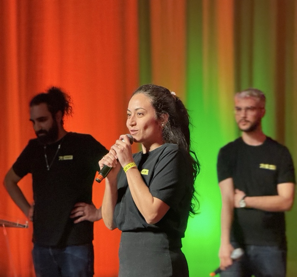 Hend Fellah speaking in front of the attendees. We can see Yoann Fleury, and Aziz Ouertani behind her